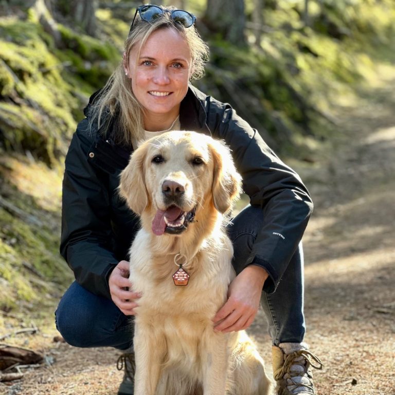 Frau mit blonden Haaren kniet neben einem lächelnden Golden Retriever auf einem Waldweg.